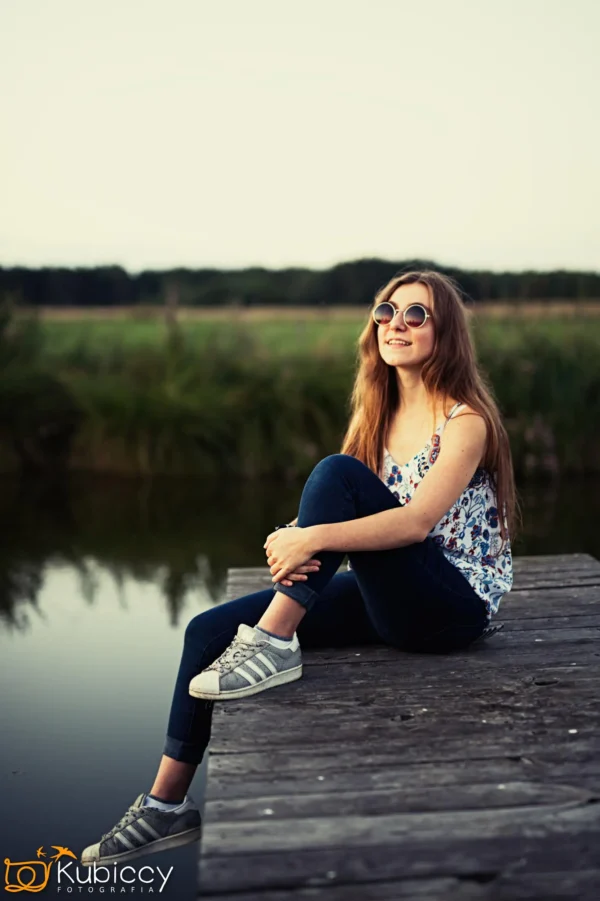 Młoda kobieta z długimi włosami, ubrana w okulary przeciwsłoneczne, kwiecisty top, dżinsy i trampki, siedzi uśmiechnięta na drewnianej przystani nad spokojnym zbiornikiem wodnym z trawiastymi polami w tle. - Kubiccy Fotografia - Fotograf Łódź
