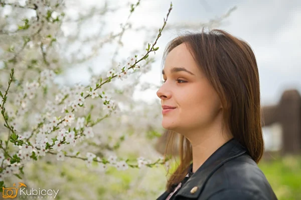 Młoda kobieta z prostymi brązowymi włosami i czarną skórzaną kurtką stoi z profilu w pobliżu kwitnących białych kwiatów na gałęzi drzewa, z miękkim tłem zieleni i nieba. - Kubiccy Fotografia - Fotograf Łódź