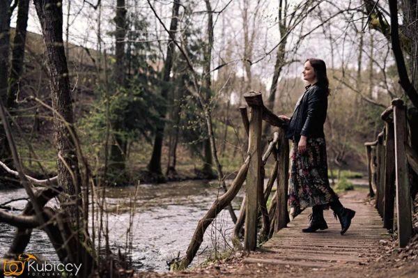 Kobieta w kwiecistej spódnicy i czarnym żakiecie stoi na rustykalnym drewnianym moście nad strumieniem, otoczonym nagimi drzewami w lesie. Patrzy przed siebie, uśmiechając się, z jedną ręką opartą na poręczy mostu. - Kubiccy Fotografia - Fotograf Łódź