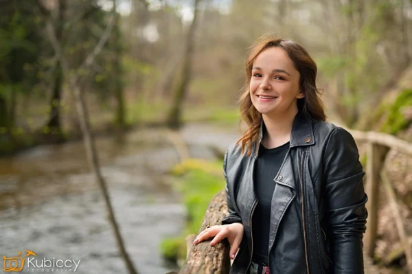 Młoda kobieta z długimi brązowymi włosami, ubrana w czarną skórzaną kurtkę, uśmiecha się, stojąc przy drewnianej balustradzie obok rzeki w zalesionym obszarze. Tło jest rozmyte drzewami i zielenią. - Kubiccy Fotografia - Fotograf Łódź