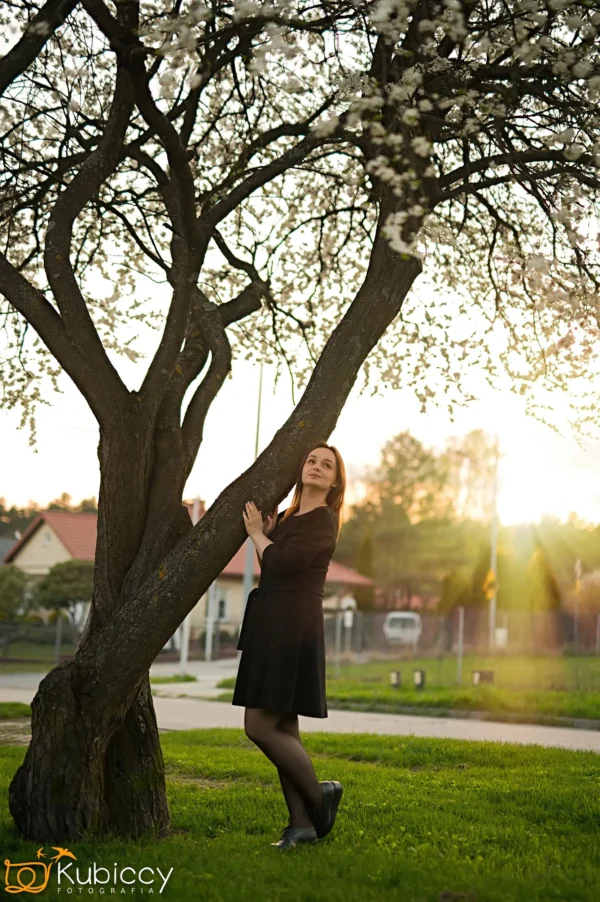 Kobieta w czarnej sukience stoi pod kwitnącym drzewem o zachodzie słońca, delikatnie trzymając gałąź. W tle widać domy, zieleń i delikatne promienie słońca. - Kubiccy Fotografia - Fotograf Łódź