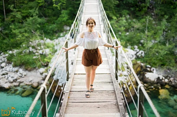 Uśmiechnięta młoda kobieta w białym topie i brązowych szortach idzie pewnie przez drewniany wiszący most nad turkusową rzeką, otoczoną bujnymi zielonymi drzewami i skałami. - Kubiccy Fotografia - Fotograf Łódź