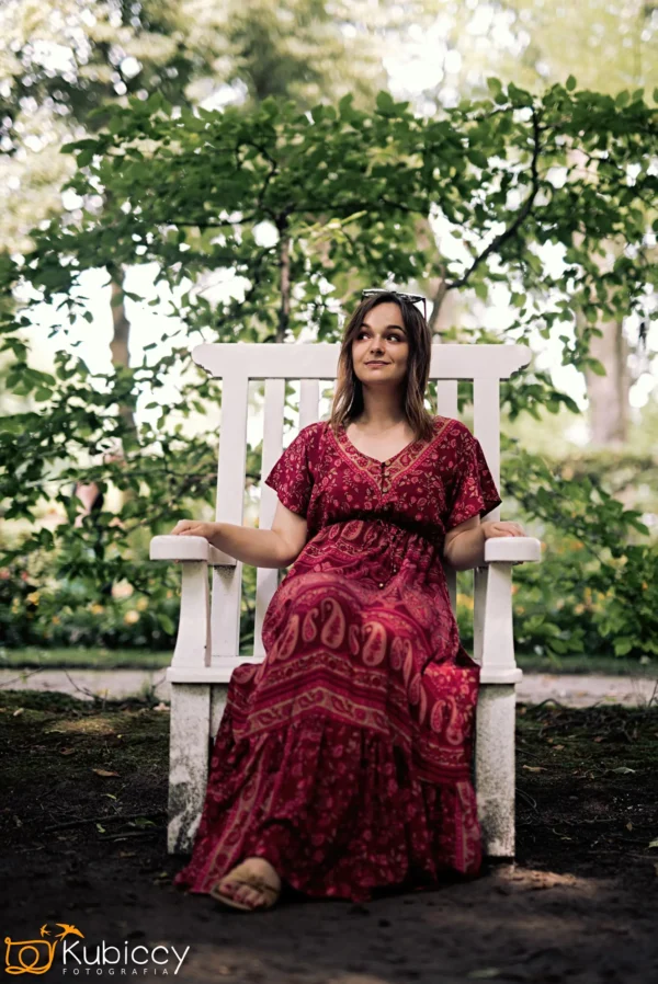 Kobieta w długiej, wzorzystej czerwonej sukni siedzi na białym drewnianym krześle na zewnątrz, otoczona bujnymi zielonymi drzewami. Spogląda lekko w górę i na boki, z rozluźnionym wyrazem twarzy. - Kubiccy Fotografia - Fotograf Łódź
