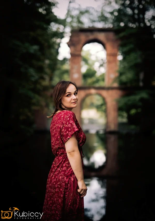 Kobieta w czerwonej wzorzystej sukience stoi nad spokojnym zbiornikiem wodnym, spoglądając przez ramię. Za nią znajduje się stary ceglany most łukowy otoczony bujnymi zielonymi drzewami. - Kubiccy Fotografia - Fotograf Łódź