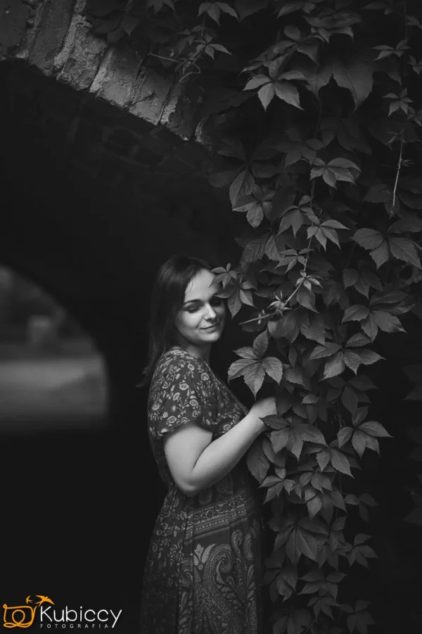 Kobieta we wzorzystej sukience stoi pod łukowatą ceglaną konstrukcją, delikatnie dotykając liściastych pnączy. Uśmiecha się delikatnie z zamkniętymi oczami. Obraz jest czarno-biały. - Kubiccy Fotografia - Fotograf Łódź