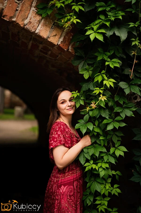 Kobieta w czerwonej wzorzystej sukience stoi pod ceglanym łukiem, uśmiechając się i trzymając liściastą zieloną winorośl, która wspina się po ścianie obok niej. Tło jest delikatnie rozmyte. - Kubiccy Fotografia - Fotograf Łódź
