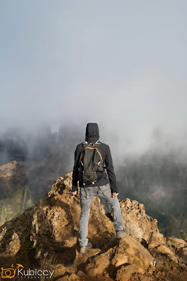 Osoba w czarnej kurtce i plecaku stoi na skalistym szczycie górskim, zwrócona plecami do kamery w kierunku mglistych chmur i nierównego terenu. - Kubiccy Fotografia - Fotograf Łódź