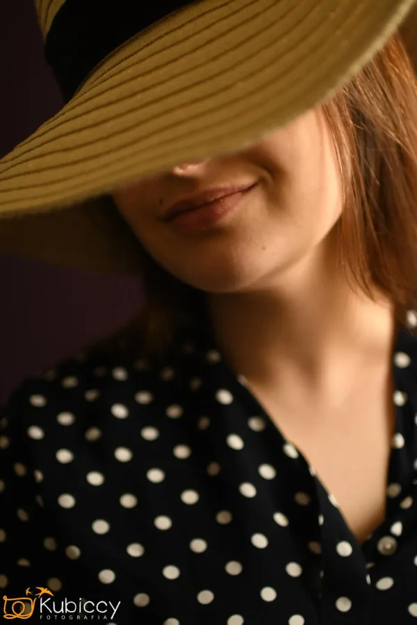 Kobieta w słomkowym kapeluszu z szerokim rondem, który częściowo zakrywa jej twarz, uśmiecha się delikatnie. Ma brązowe włosy i jest ubrana w czarną koszulę w białe kropki. Tło jest ciemne i nieostre. - Kubiccy Fotografia - Fotograf Łódź