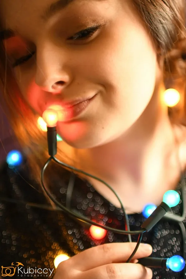 Młoda kobieta o jasnej karnacji uśmiecha się delikatnie, trzymając kolorowe, świecące lampki na sznurku blisko twarzy. Światła oświetlają jej rysy, a ona ma na sobie błyszczący czarny top. - Kubiccy Fotografia - Fotograf Łódź