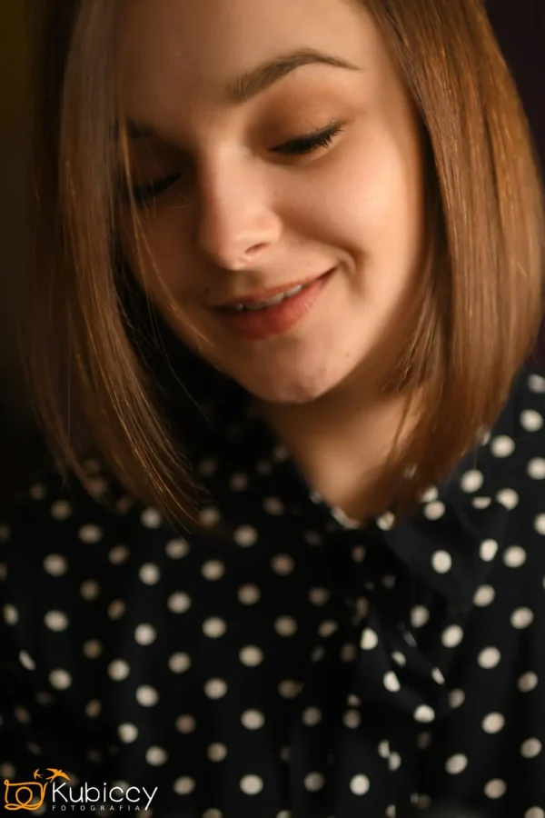 Kobieta z prostymi, sięgającymi ramion brązowymi włosami uśmiecha się delikatnie, patrząc w dół. Ma na sobie granatową bluzkę w białe kropki. Zdjęcie ma ciepłą, miękką ostrość. - Kubiccy Fotografia - Fotograf Łódź