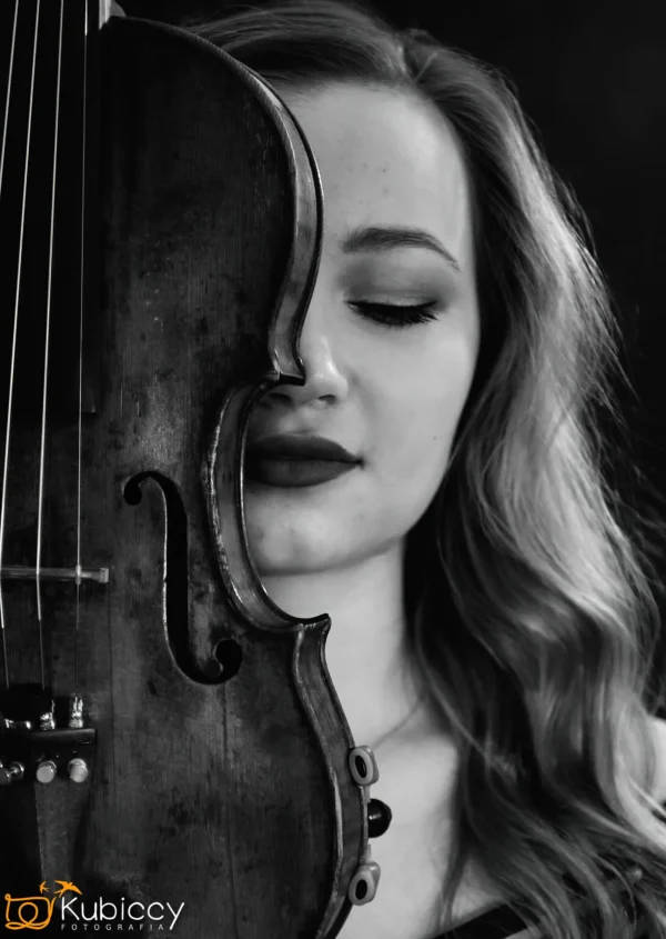 Kobieta z falującymi włosami pozuje z zamkniętymi oczami, trzymając skrzypce pionowo, tak aby zakrywały połowę jej twarzy. Zdjęcie jest czarno-białe, tworząc dramatyczny i artystyczny efekt. - Kubiccy Fotografia - Fotograf Łódź