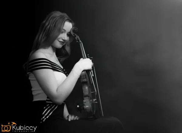 Młoda kobieta z długimi włosami siedzi z profilu, uśmiechając się delikatnie i trzymając skrzypce w pozycji pionowej. Ma na sobie sukienkę z odkrytymi ramionami w paski. Zdjęcie ma miękki, czarno-biały efekt z ciemnym tłem. - Kubiccy Fotografia - Fotograf Łódź