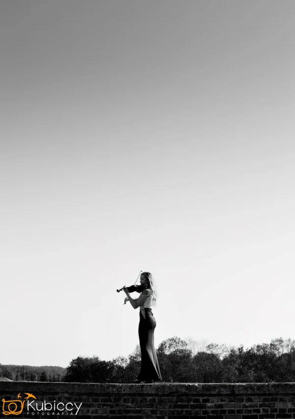 Kobieta stoi na zewnątrz na gzymsie grając na skrzypcach, z drzewami w tle i rozległym, czystym niebem powyżej. Obraz jest czarno-biały. - Kubiccy Fotografia - Fotograf Łódź