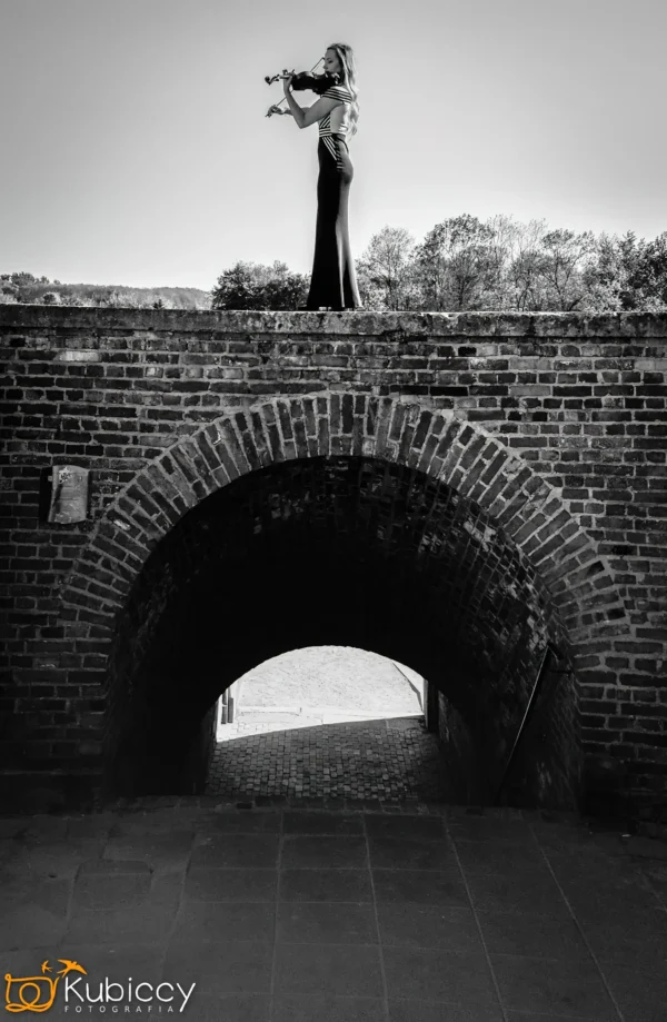 Kobieta w długiej sukni gra na skrzypcach, stojąc na szczycie ceglanego mostu łukowego, z drzewami i czystym niebem w tle. Obraz jest czarno-biały. - Kubiccy Fotografia - Fotograf Łódź