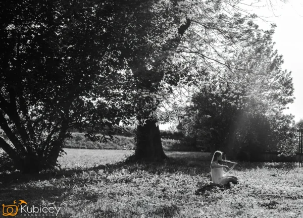 Młoda dziewczyna siedzi i gra na skrzypcach na nasłonecznionym trawiastym obszarze pod dużymi drzewami, gdzie otacza ją pstrokate światło i cienie. Atmosfera jest spokojna i pogodna. - Kubiccy Fotografia - Fotograf Łódź