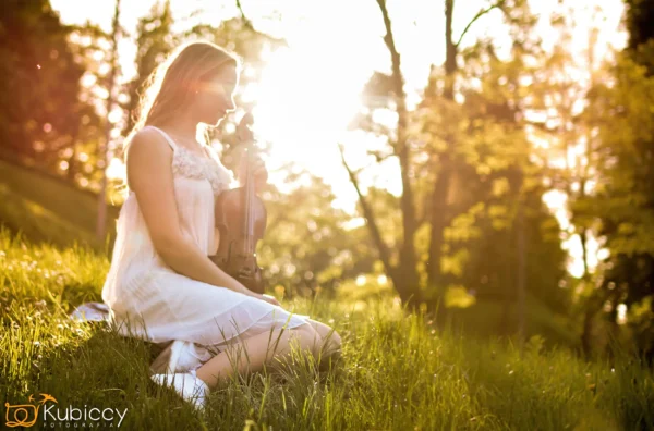 Kobieta w białej sukience siedzi na trawie w oświetlonym słońcem parku, trzymając skrzypce. Tło jest jasne i rozmyte, z drzewami i promieniami słońca. - Kubiccy Fotografia - Fotograf Łódź