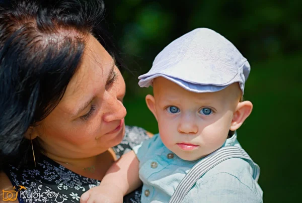 Kobieta o ciemnych włosach uśmiecha się, trzymając dziecko o niebieskich oczach w jasnej czapce i koszuli. Dziecko patrzy bezpośrednio w kamerę na rozmytym zielonym tle. - Kubiccy Fotografia - Fotograf Łódź