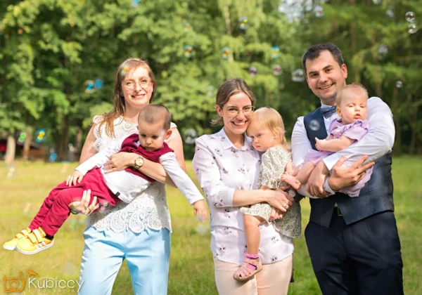 Troje dorosłych stoi na zewnątrz, trzymając trójkę małych dzieci, uśmiechając się do kamery w słoneczny dzień. Tło wypełniają drzewa i trawa, a w powietrzu unoszą się bąbelki. - Kubiccy Fotografia - Fotograf Łódź