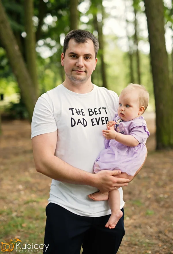 Mężczyzna w białej koszulce z napisem The Best Dad Ever trzyma dziecko w fioletowej sukience, stojąc na zewnątrz w zalesionym obszarze. Oboje wyglądają na spokojnych i zadowolonych. - Kubiccy Fotografia - Fotograf Łódź