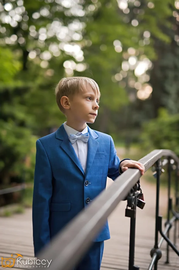 Młody chłopak w niebieskim garniturze i muszce stoi na moście, trzymając się poręczy i patrząc w zamyśleniu w bok, na rozmytym tle zielonych drzew. - Kubiccy Fotografia - Fotograf Łódź