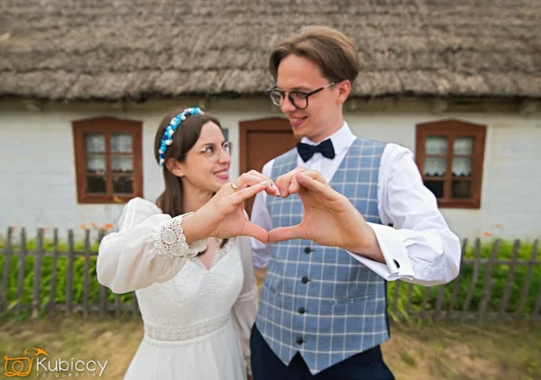 Panna młoda i pan młody stoją przed rustykalnym domem, uśmiechając się do siebie i tworząc kształt serca rękami. Panna młoda ma na sobie białą sukienkę i opaskę, a pan młody kamizelkę, muszkę i okulary. - Kubiccy Fotografia - Fotograf Łódź