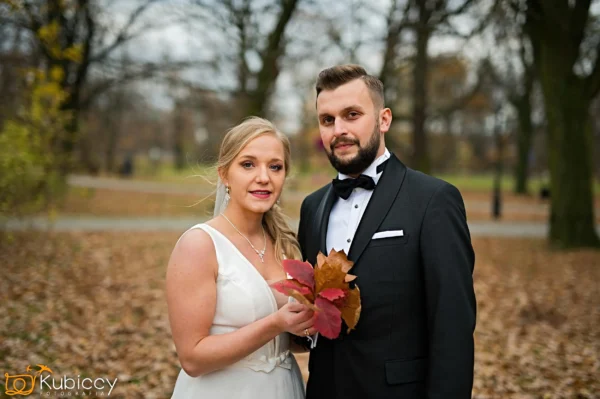 Panna młoda w białej sukni trzyma jesienne liście, stojąc obok pana młodego w czarnym smokingu. Są na zewnątrz w parku z opadłymi liśćmi i nagimi drzewami w tle. - Kubiccy Fotografia - Fotograf Łódź