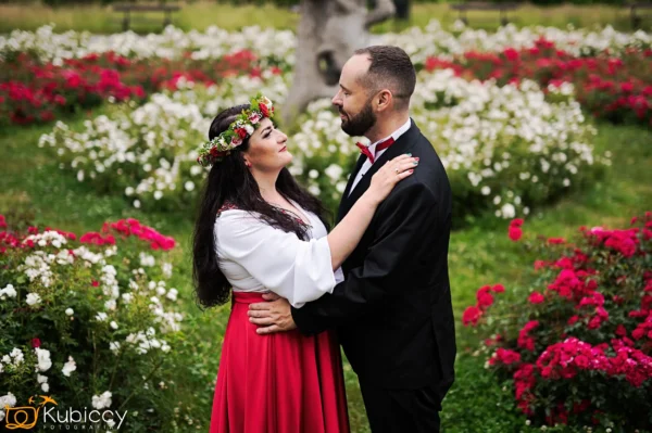 Para ubrana w tradycyjne stroje obejmuje się w ogrodzie otoczonym kwitnącymi czerwonymi i białymi kwiatami. Kobieta ma na sobie kwiecistą koronę i czerwoną spódnicę, podczas gdy mężczyzna jest ubrany w czarny garnitur z czerwoną muszką. - Kubiccy Fotografia - Fotograf Łódź