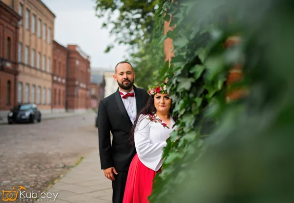 Mężczyzna w czarnym smokingu i kobieta w białej bluzce i czerwonej spódnicy z kwiatową koroną stoją na brukowanej ulicy, częściowo ukrytej przez zielony bluszcz, z ceglanymi budynkami w tle. - Kubiccy Fotografia - Fotograf Łódź