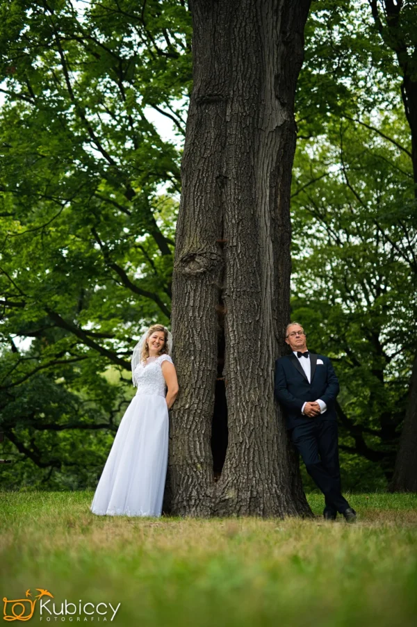 Mężczyzna i kobieta w strojach ślubnych opierający się o drzewo. - Kubiccy Fotografia - Fotograf Łódź