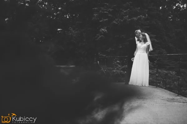 Czarno-białe zdjęcie panny młodej i pana młodego stojących blisko siebie na ścieżce w lesie, dzielących czułą chwilę. Panna młoda ma na sobie długą suknię i welon, a pan młody pochyla się, by ją pocałować. - Kubiccy Fotografia - Fotograf Łódź