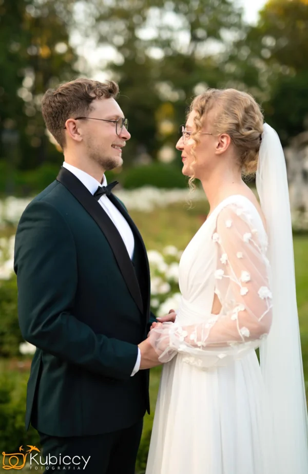 Panna młoda i pan młody stoją naprzeciwko siebie na zewnątrz, trzymając się za ręce i uśmiechając się. Pan młody ma na sobie ciemny garnitur i muszkę; panna młoda ma na sobie białą sukienkę z przezroczystymi rękawami i welon. W tle zieleń i kwiaty. - Kubiccy Fotografia - Fotograf Łódź