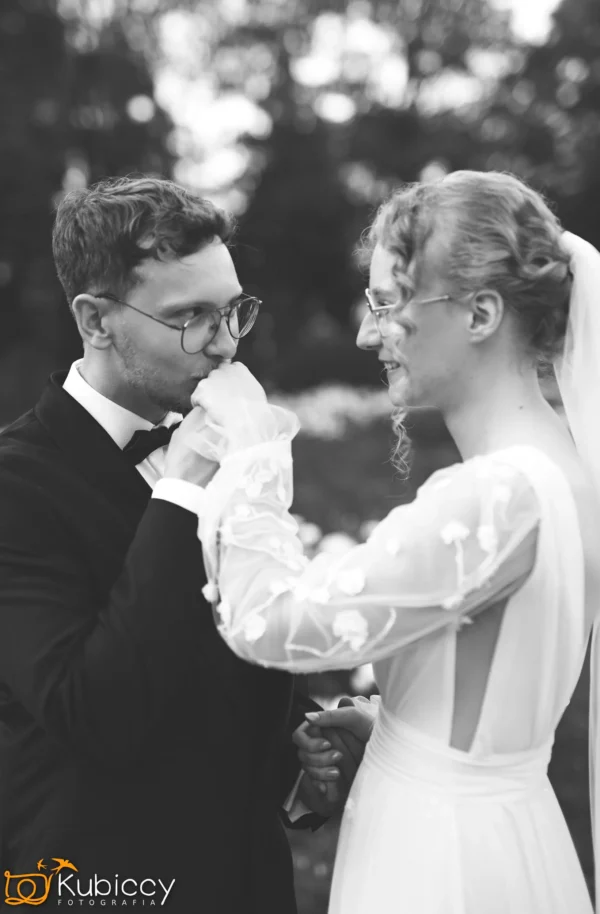 Pan młody w garniturze delikatnie całuje rękę panny młodej, która uśmiecha się, oboje ubrani formalnie. Panna młoda ma na sobie haftowaną suknię z długimi rękawami i welon. Zdjęcie jest czarno-białe, z rozmytym tłem na zewnątrz. - Kubiccy Fotografia - Fotograf Łódź