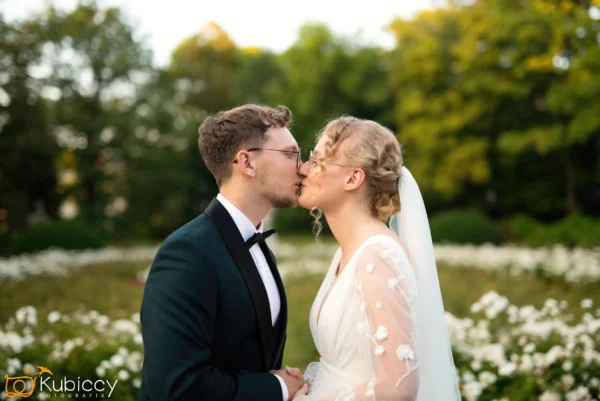 Panna młoda i pan młody stoją na zewnątrz w ogrodzie, ubrani w stroje ślubne, delikatnie całując się w usta z kwiatami i zielonymi drzewami w tle. - Kubiccy Fotografia - Fotograf Łódź