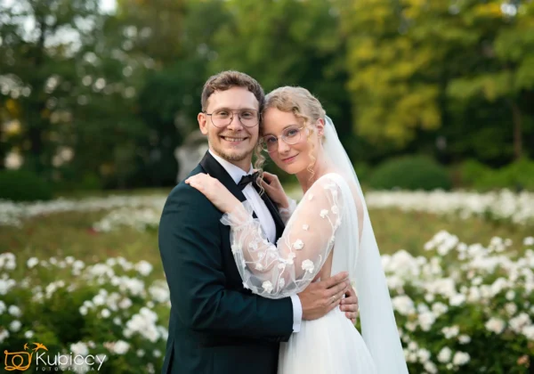 Panna młoda i pan młody uśmiechają się, obejmując się na zewnątrz, otoczeni kwitnącymi białymi kwiatami i zielenią. Oboje noszą okulary; panna młoda ma białą suknię i welon, a pan młody czarny garnitur i muszkę. - Kubiccy Fotografia - Fotograf Łódź