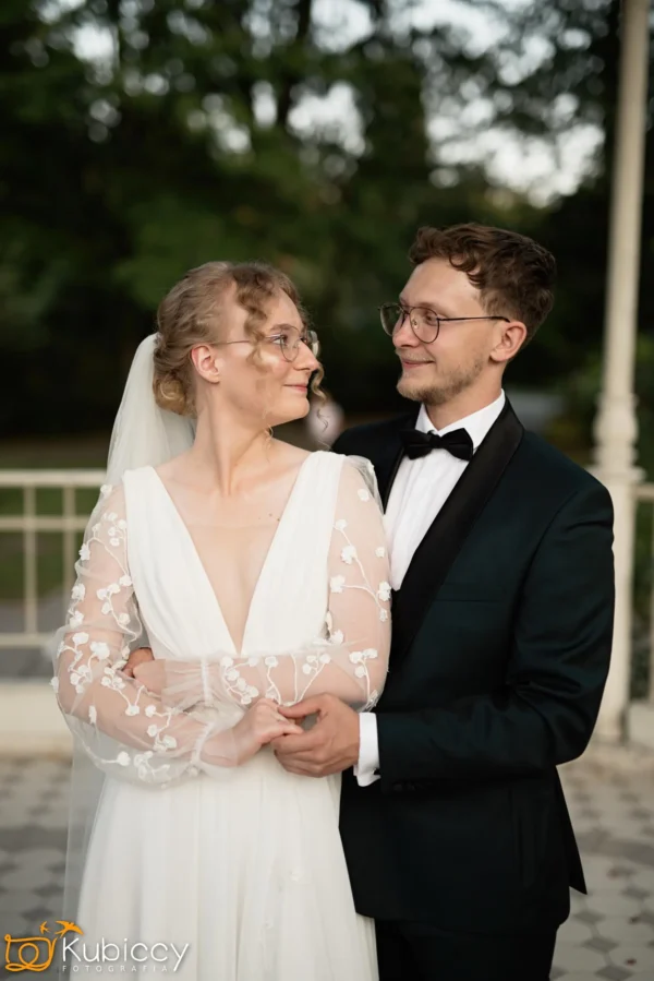Panna młoda i pan młody stoją blisko siebie, uśmiechając się i patrząc sobie w oczy. Panna młoda ma na sobie białą suknię z przezroczystymi, kwiecistymi rękawami i welonem; pan młody ma na sobie czarny smoking i okulary. Są na zewnątrz w pobliżu balustrady. - Kubiccy Fotografia - Fotograf Łódź