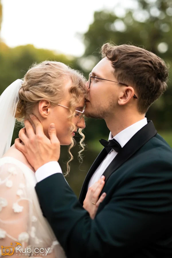 Pan młody w czarnym smokingu delikatnie całuje pannę młodą w czoło, trzymając ją za twarz. Panna młoda, w okularach, welonie i haftowanej sukni, stoi blisko z ręką na jego piersi. Są na zewnątrz, otoczeni zielenią. - Kubiccy Fotografia - Fotograf Łódź