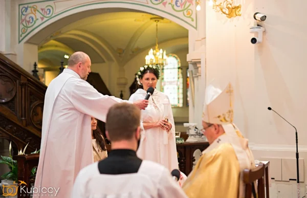 Kobieta w białej szacie mówi do mikrofonu trzymanego przez księdza podczas ceremonii kościelnej, z innymi duchownymi siedzącymi w pobliżu i witrażami w tle. - Kubiccy Fotografia - Fotograf Łódź