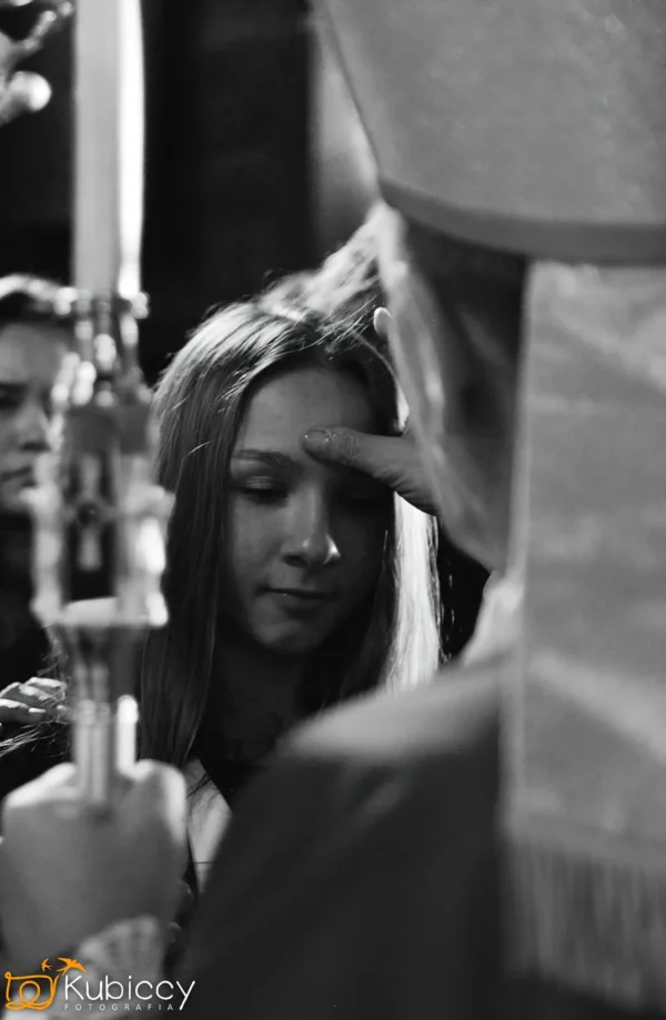 Młoda dziewczyna z zamkniętymi oczami otrzymuje błogosławieństwo na czole od duchownego podczas ceremonii religijnej. Inne osoby stoją w pobliżu. Obraz jest czarno-biały. - Kubiccy Fotografia - Fotograf Łódź