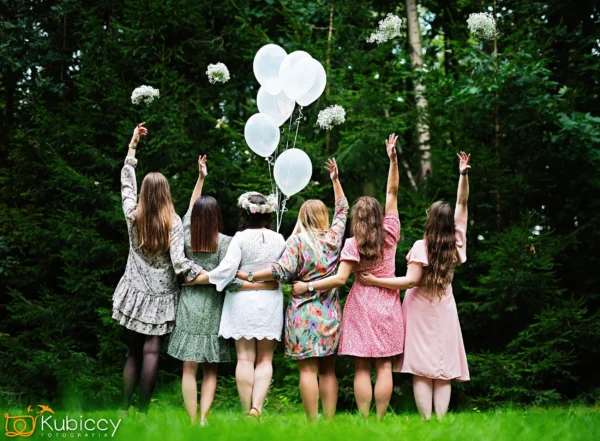 Sześć kobiet w kolorowych sukienkach stoi ramię w ramię, plecami do kamery, unosząc ręce i trzymając białe balony i kwiaty na tle zielonych drzew. - Kubiccy Fotografia - Fotograf Łódź