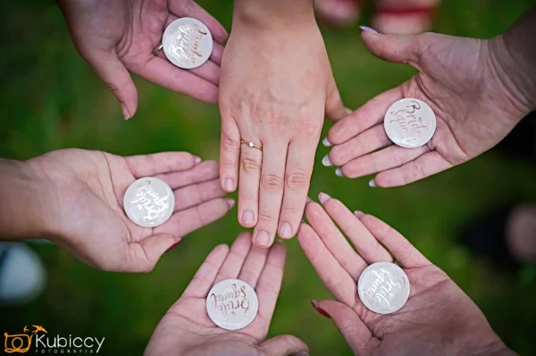 Sześć rąk ułożonych w okrąg trzyma przyciski "Bride Squad", podczas gdy jedna ręka z pierścionkiem zaręczynowym spoczywa pośrodku nad trawą. Znak wodny fotografa jest widoczny w rogu. - Kubiccy Fotografia - Fotograf Łódź