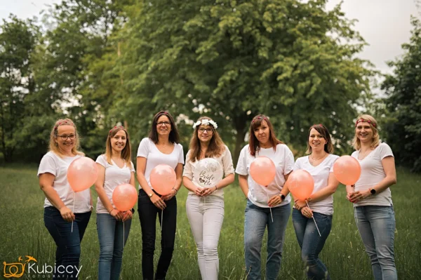 Siedem kobiet stoi razem na trawie, wszystkie ubrane w białe topy i dżinsy. Kobieta w centrum trzyma balon Panny Młodej i nosi kwiatową koronę, podczas gdy inne trzymają różowe balony. Tło wypełniają drzewa. - Kubiccy Fotografia - Fotograf Łódź