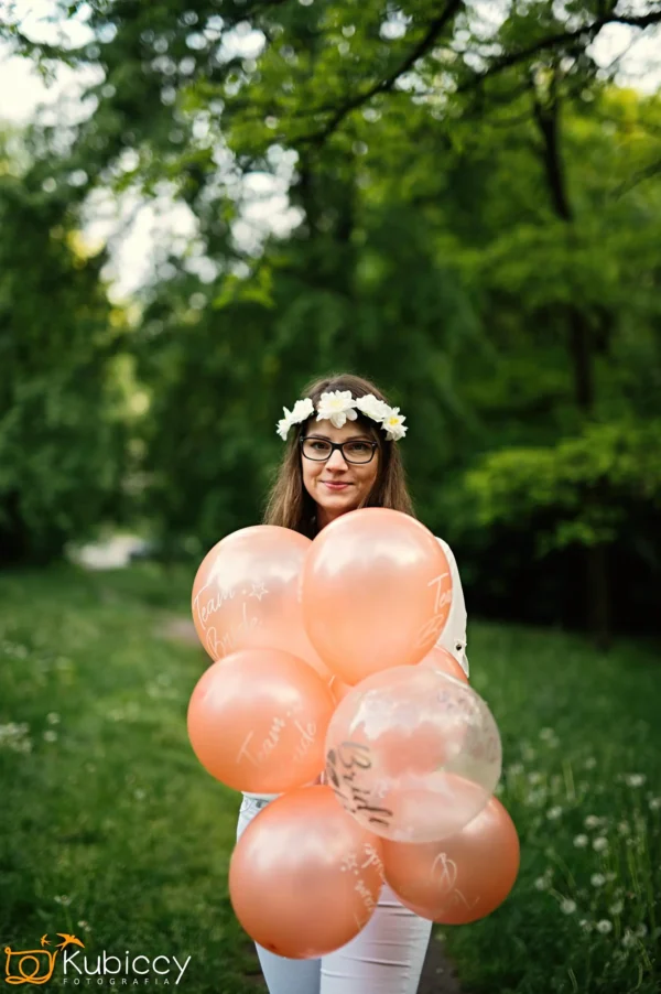 Kobieta w okularach i koronie z kwiatów trzyma kilka brzoskwiniowych balonów "Team Bride", stojąc na ścieżce w zielonym, zalesionym parku. - Kubiccy Fotografia - Fotograf Łódź