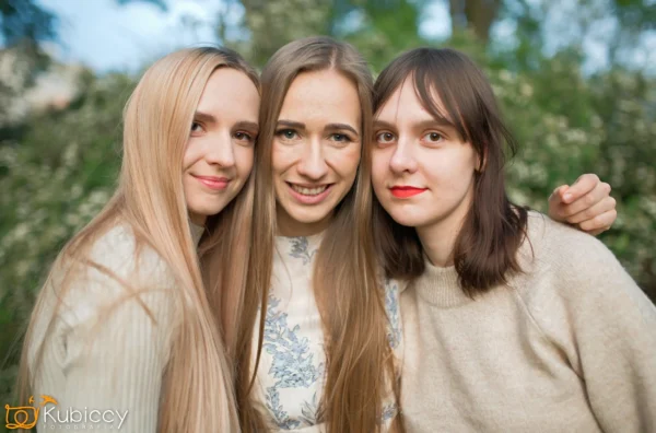 Trzy młode kobiety z długimi włosami, ubrane w jasne swetry, stoją blisko siebie na zewnątrz, uśmiechając się do kamery z zielenią w rozmytym tle. - Kubiccy Fotografia - Fotograf Łódź