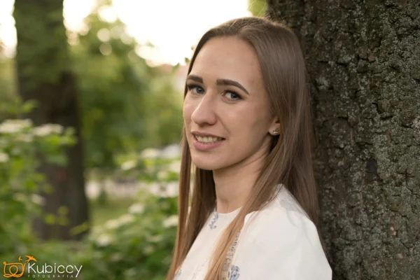 Kobieta z prostymi jasnobrązowymi włosami i białą bluzką uśmiecha się lekko, opierając się o drzewo w zielonej scenerii na zewnątrz. Tło jest delikatnie rozmyte z widocznymi liśćmi i światłem słonecznym. - Kubiccy Fotografia - Fotograf Łódź