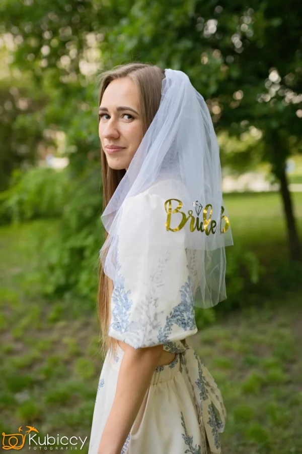 Młoda kobieta z długimi brązowymi włosami ma na sobie białą sukienkę z niebieskimi wzorami i biały welon ze złotym napisem Bride. Stoi na zewnątrz w zielonym, pełnym liści parku, spoglądając przez ramię i uśmiechając się delikatnie. - Kubiccy Fotografia - Fotograf Łódź