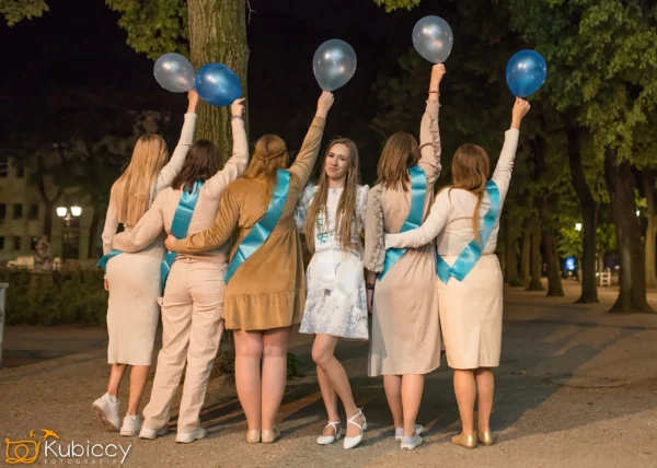Sześć kobiet stoi na zewnątrz w nocy, pięć z nich tyłem do kamery w beżowych strojach, niebieskich szarfach i trzymających niebieskie balony, podczas gdy jedna jest uśmiechnięta, ubrana w białą sukienkę i niebieskie buty. W tle widać drzewa. - Kubiccy Fotografia - Fotograf Łódź
