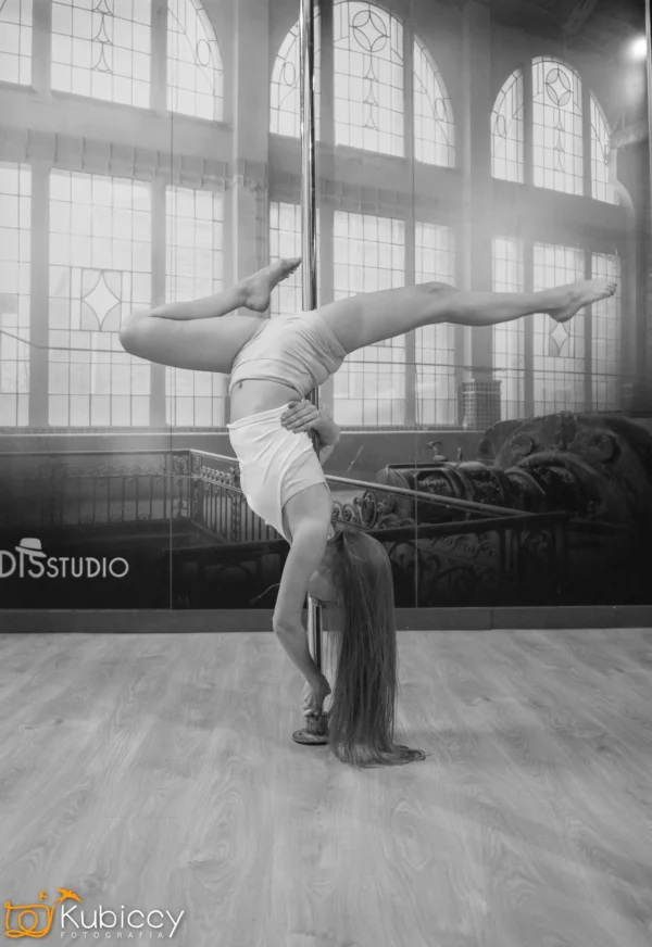 Osoba wykonuje zaawansowany ruch pole dance, balansując do góry nogami na rurze z jedną nogą zaczepioną, a drugą wyciągniętą poziomo, wewnątrz studia z dużymi łukowatymi oknami. - Kubiccy Fotografia - Fotograf Łódź