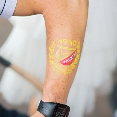 Ramię osoby z namalowanym czerwono-żółtym emblematem Widzewa 1910, otoczonym wieńcem laurowym. Osoba ma na sobie czarny smartwatch i białą koszulkę z krótkim rękawem. - Kubiccy Fotografia - Fotograf Łódź