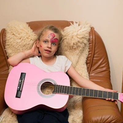 Młoda dziewczyna z pomalowaną twarzą różowego flaminga relaksuje się na opalonym fotelu, trzymając na kolanach różową gitarę akustyczną. Patrzy w kamerę, opierając rękę na krześle. - Kubiccy Fotografia - Fotograf Łódź