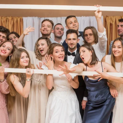 Grupa ludzi w formalnych strojach wesoło pozuje razem na weselu, trzymając przed sobą dużą białą ramę ozdobioną kwiatami. Para młoda stoi w centrum, otoczona przez uśmiechniętych przyjaciół. - Kubiccy Fotografia - Fotograf Łódź